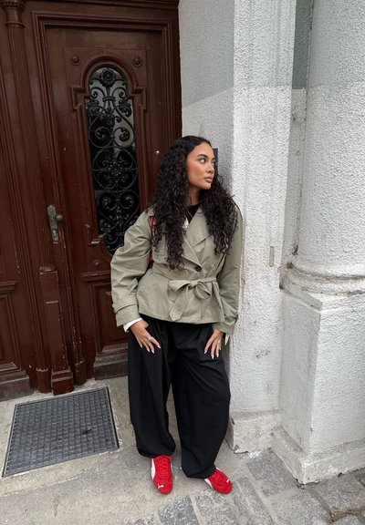 Chaqueta verde oliva con cinturón, pantalones negros oversize y zapatillas rojas. Cabello rizado con un brillo sutil, de pie cerca de una puerta de madera.