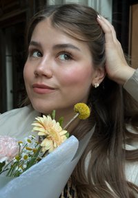 Kvinne med langt brunt hår iført en beige frakk holder en bukett med gule og rosa blomster, pakket inn i hvitt papir. Nærbilde, nøytral bakgrunn.