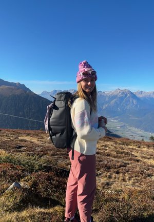 Mochila de material negro llevada por una mujer con un suéter crema que presenta patrones en tonos pastel y pantalones rosas, de pie en un terreno montañoso.