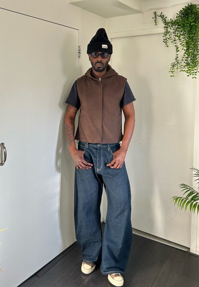 Hombre con gorro negro, gafas de sol oscuras, chaleco marrón sin mangas con cremallera, camiseta negra, vaqueros azules de pierna ancha y zapatos blancos de pie dentro cerca de plantas.