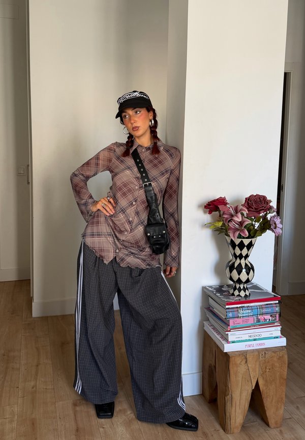 Una mujer posa en un interior con gorra, camisa de cuadros, pantalones de pernera ancha y bolso bandolera, junto a un jarrón con flores sobre una pila de libros.