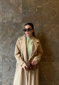 Bež plašč trench, zelen pulover, črna sončna očala, drži rjavo tkan torbico. Gladki, strukturirani materiali s prilagojeno prileganjem.