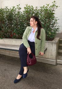 Vrouw in een groene jas en marineblauwe broek die op een bankje zit, met een geweven bordeauxrode handtas in haar hand, omringd door groene struiken en een betonnen muur op de achtergrond.