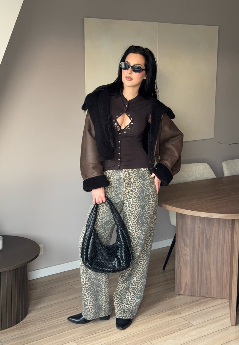 Femme portant des lunettes de soleil, une veste en peau lainée brune, un haut noir découpé, un pantalon à imprimé léopard, tenant un sac tissé noir, debout à l'intérieur près d'une table.
