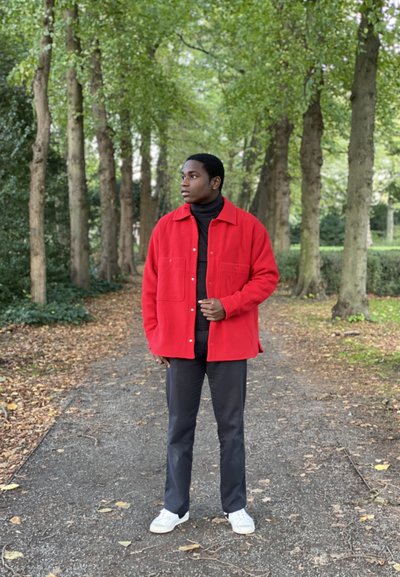 Camisa chaqueta roja de botón, hecha de material suave, con dos bolsillos frontales, llevada sobre un cuello de tortuga negro y combinada con pantalones negros y zapatillas deportivas blancas.