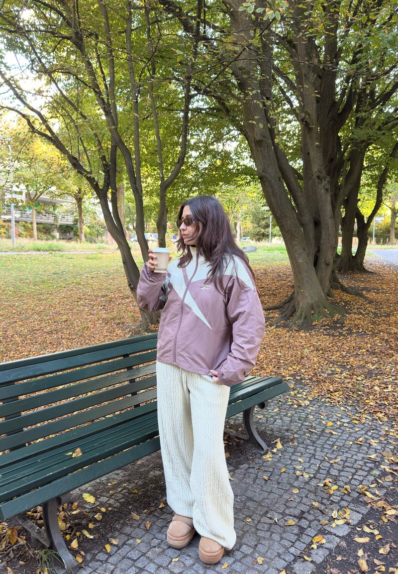 Une femme portant des lunettes de soleil tient un gobelet à emporter à côté d’un banc de parc, sous des arbres aux couleurs automnales. Des feuilles mortes couvrent le chemin.