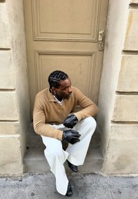 Homme aux cheveux tressés portant des gants noirs, un pull beige, un pantalon blanc et des chaussures noires assis sur des marches en pierre devant une porte beige.