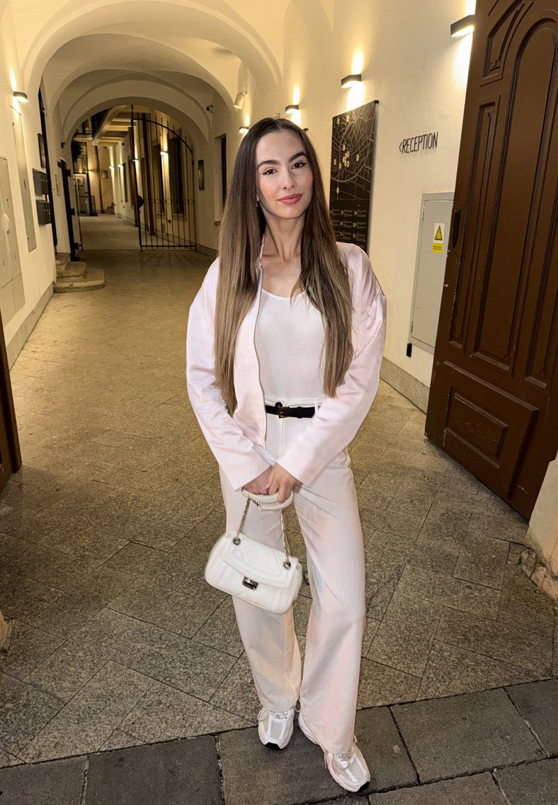 Jeune femme debout dans un couloir voûté bien éclairé, vêtue d'un costume rose clair, d'un haut blanc et de baskets, tenant un petit sac blanc devant un panneau de réception.