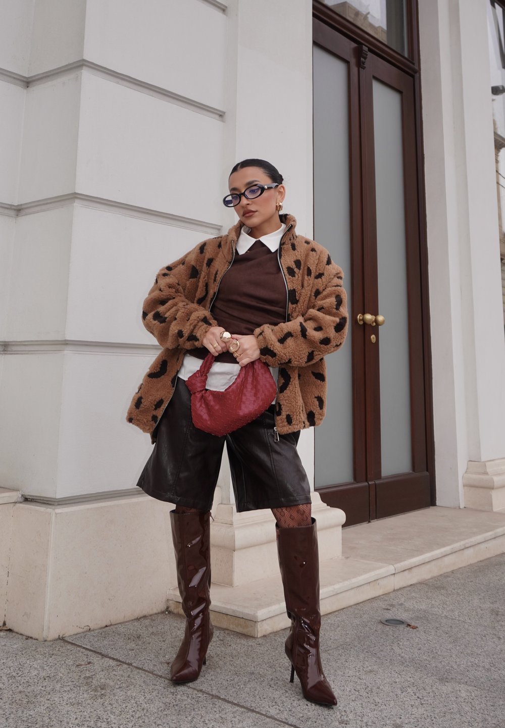 Veste marron duveteuse avec des taches noires, pull marron, shorts en cuir noir, bottes brillantes marron jusqu'aux genoux, et sac à main rouge texturé.