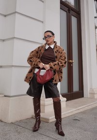 Veste marron duveteuse avec des taches noires, pull marron, shorts en cuir noir, bottes brillantes marron jusqu'aux genoux, et sac à main rouge texturé.