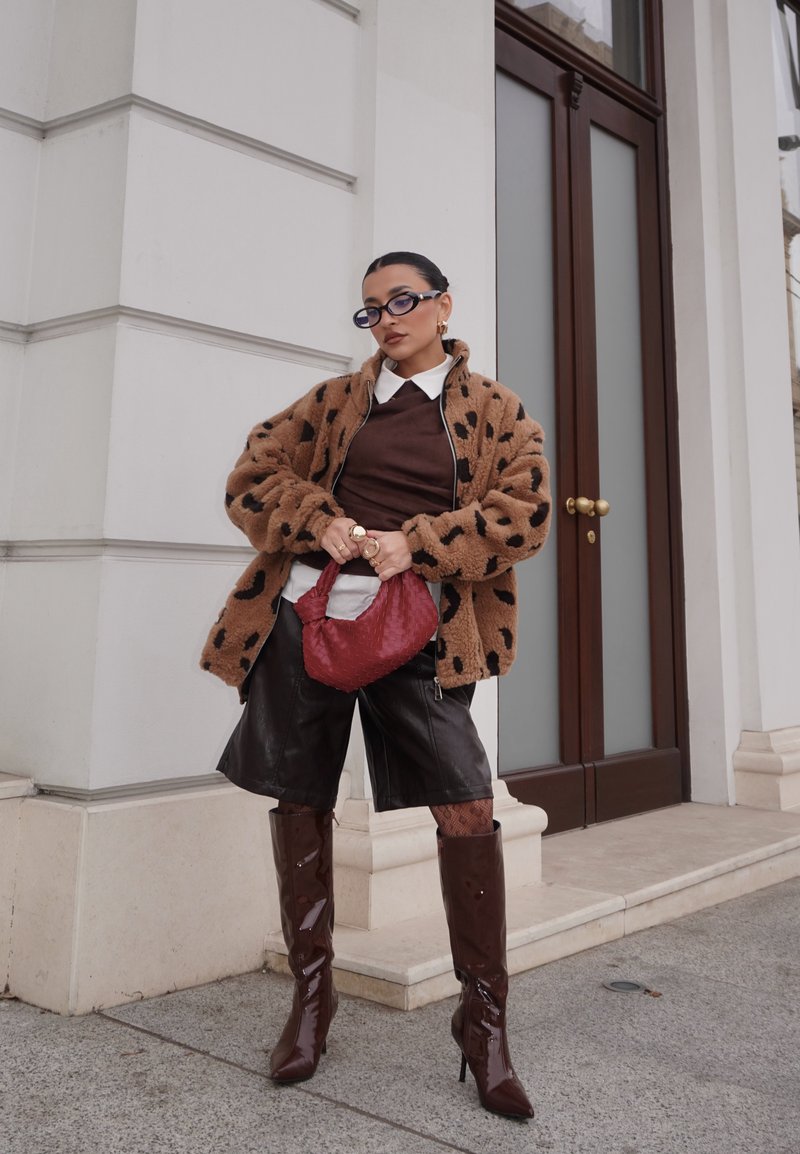Veste marron duveteuse avec des taches noires, pull marron, shorts en cuir noir, bottes brillantes marron jusqu'aux genoux, et sac à main rouge texturé.