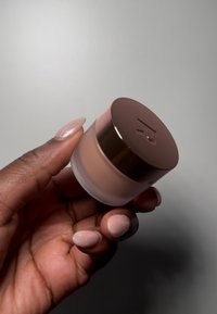 Gjennomsiktig krukke med et produkt i blush rosa farge, toppet med et blankt brunt lokk med en preget logo. Hånd som holder krukken.