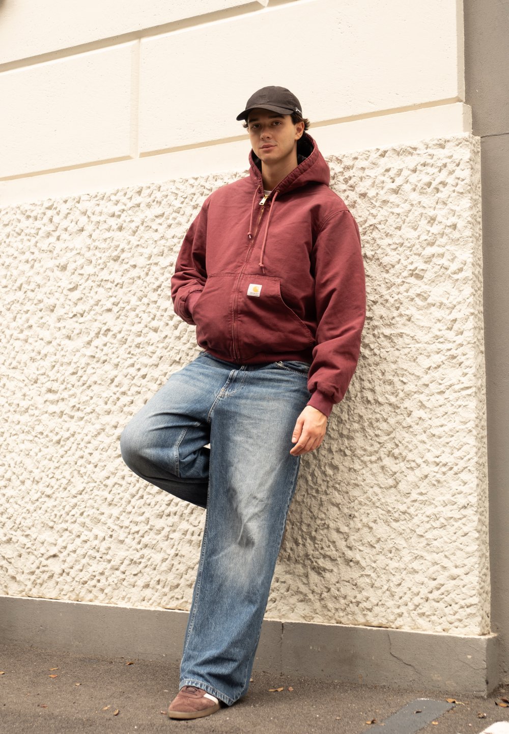 Μαροτίνα φούτερ με φερμουάρ, ανοιχτό μπλε φαρδιά τζιν, καφέ παπούτσια. Άτομο στηρίζεται σε υφασμένη άσπρη κουρτίνα, φοράει μαύρο καπέλο. Καθημ�ερινό ντύσιμο.