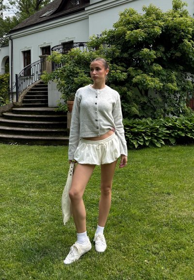 Cárdigan de punto gris claro con botones, pantalones cortos blancos con fruncidos y zapatillas blancas. De pie sobre la hierba cerca de un edificio y la vegetación.