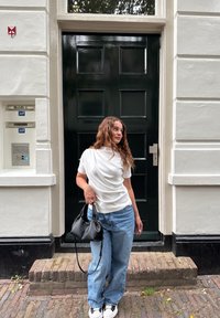 Hvit drapert t-skjorte, løse lyseblå jeans, svart veske, og hvite joggesko. Svart dør med rektangulære paneler og grå murtrapper.