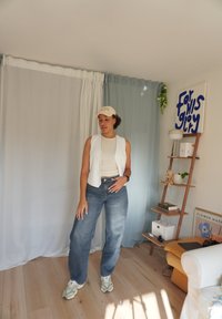 Biely bezrukávový sveter na béžovom topi, spárovaný s svetlomodrými širokými džínsami a sivými teniskami. Pozadie tvorí svetlé závesy a dekorácie.