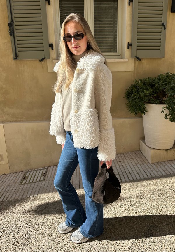 Krémový fleecový kabát s texturovanou látkou a velkými knoflíky, zkombinovaný s modrými zvonovými džíny a šedými teniskami. V ruce černá kabelka.