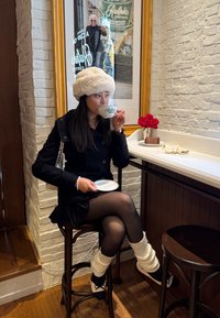 Vrouw in zwarte jas en witte bontmuts die koffie drinkt aan een cafébalie met rode rozen en een ingelijste poster aan een bakstenen muur.