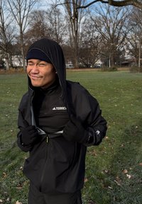 Crna vanjska jakna s kapuljačom, ima patentni zatvarač sprijeda i elastične manžete. Majica ispod također je crna, s vidljivim logotipom.