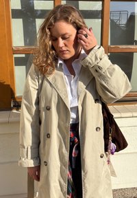 Bežinis paltas su dideliais mygtukais, plačiu apykakle ir susiaurinta juosmeniu; dėvimas ant baltos marškinėlių ir langeliais raštuotos sijono. Rudas krepšys su dekoratyvine pakabuko detalė.