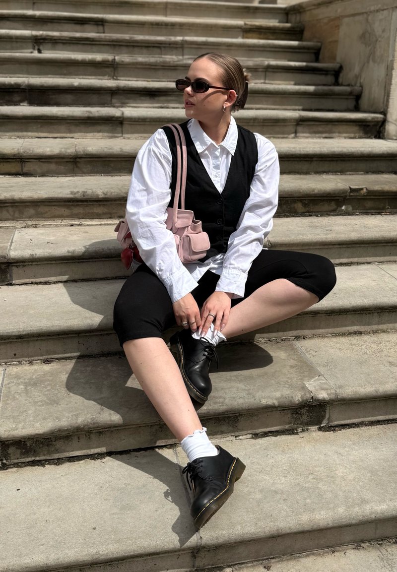 Jeune femme portant un gilet noir, une chemise blanche, un pantalon noir court et des bottes, assise sur des marches en pierre avec des lunettes de soleil et un sac rose.