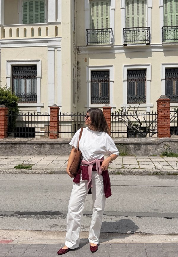 Kvinde i hvidt outfit med pink sweater bundet omkring taljen, bærende på brun taske, stående på fortov foran en beige bygning med jernbalkoner og rød murstensmur.