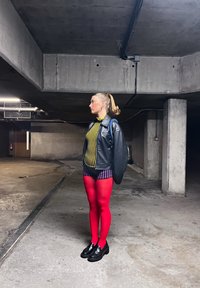 Άτομο με ξανθιά αλογοουρά στέκεται σε ένα υπόγειο γκαράζ από μπετόν φορώντας μαύρο δερμάτινο μπουφάν, μπλούζα με κίτρινη ρίγα, ροζ σορτς και φωτεινά κόκκινα καλσόν.