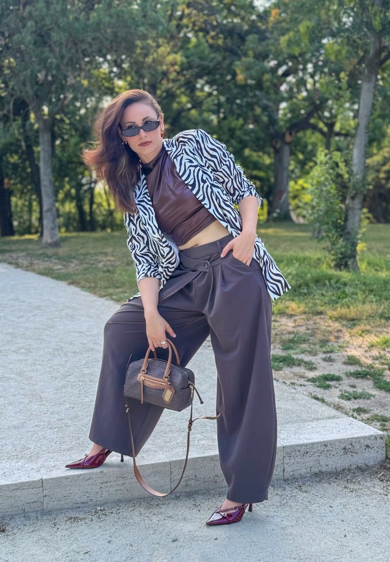 Femme portant une veste à motif zèbre, un crop top marron, un pantalon large, des talons rouges, des lunettes de soleil, tenant un sac à main, posant à l'extérieur sur un rebord en pierre.