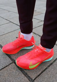 Chaussures de course Nike orange vif avec tige en mesh léger, semelle verte et accents réfléchissants, associées à des chaussettes blanches texturées.