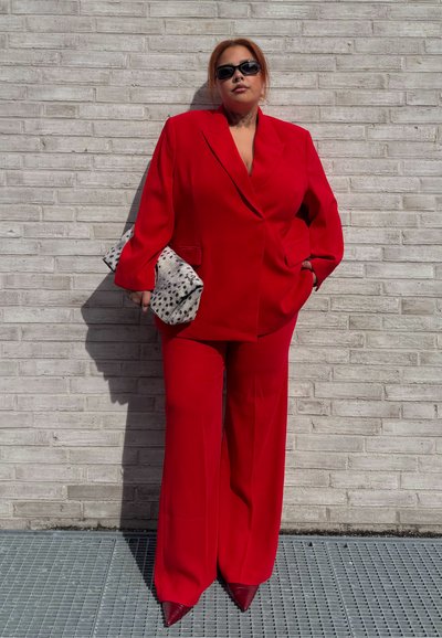 Persona con cabello naranja y gafas de sol, vestida con un traje rojo brillante y sosteniendo un bolso blanco con manchas, de pie contra una pared de ladrillos grises.