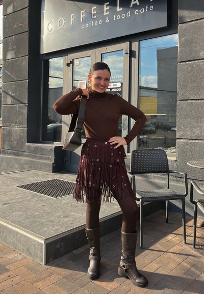 Jersey de cuello alto marrón, falda burdeos con flecos y ojetes plateados, medias negras y botas marrones hasta la rodilla. De pie frente a un café.