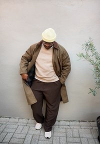 Palton lung maro peste un pulover bej și pantaloni maro cu carouri. Căciulă galben deschis și adidași albi. Suprafețe texturate și o plantă verde în apropiere.