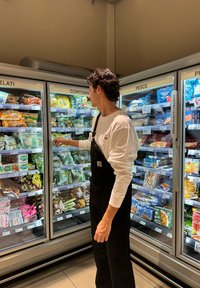 Una persona con un overol negro alcanza dentro de un congelador expositivo que contiene alimentos congelados envasados con varios colores y etiquetas.