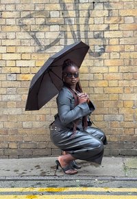 Jachetă din piele neagră și pantaloni largi, umbrelă neagră, ochelari de soare eleganți, stând pe trotuar umed lângă un zid de cărămidă galbenă texturată.
