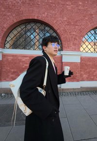 Jeune personne portant des lunettes et un manteau noir, avec un sac à dos blanc, tenant une tasse de café, marchant devant un bâtiment en briques rouges avec des fenêtres en arc.