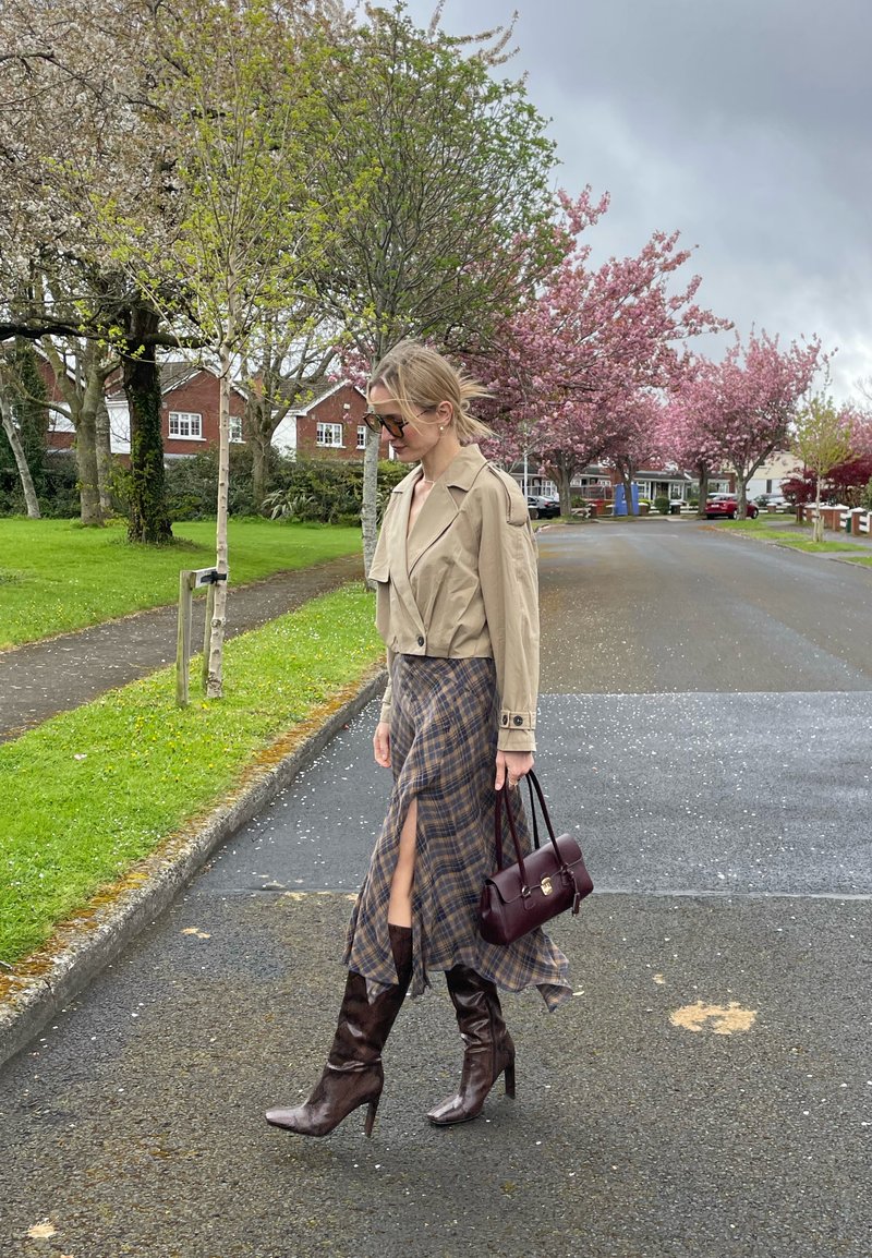 Femme portant une veste beige, une jupe à carreaux avec une fente et des bottes marron montant au genou, marchant dans une rue bordée d'arbres en fleurs et de maisons.