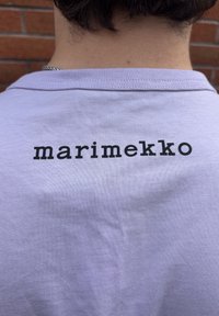 Světle fialové bavlněné tričko s potiskem "marimekko" v tučných černých písmenech na zádech, kulatým výstřihem a krátkými rukávy.