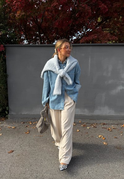 Camisa de mezclilla azul claro abotonada, combinada con un suéter gris suave atado alrededor de los hombros, pantalones anchos color crema y zapatos plateados.