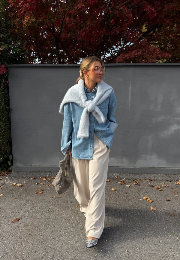 Světle modrá džínová košile na knoflíky, navrstvená s měkkým šedým svetrem přehnutým přes ramena, doplněná krémovými širokými kalhotami a stříbrnými botami.