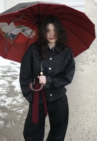 Femme aux cheveux noirs portant une tenue noire, tenant un parapluie rouge avec un motif de grue, debout sur un chemin enneigé près d'un mur en pierre.