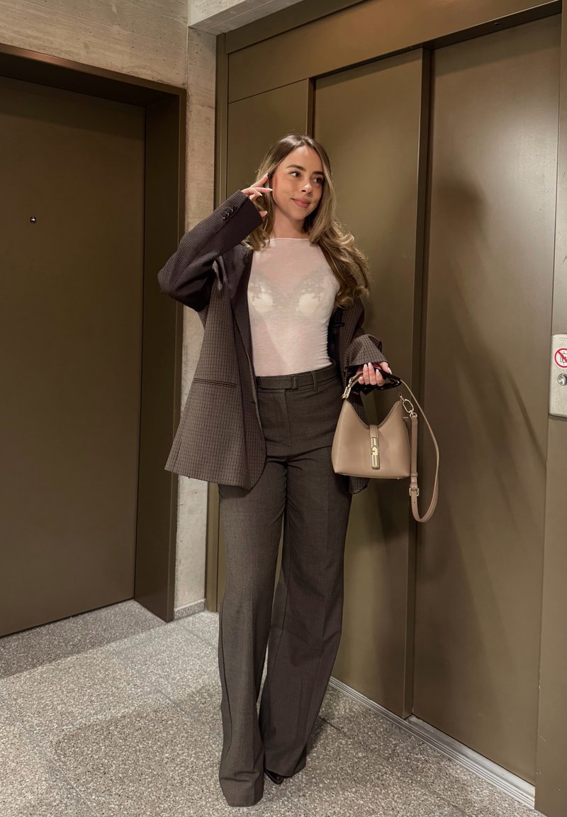 Γυναίκα με μακριά μαλλιά, ντυμένη με καφέ σακάκι και παντελόνι, κρατώντας μπεζ τσάντα, στέκεται κοντά σε κλειστές πόρτες ανελκυστήρα σε έναν διάδρομο κτιρίου.