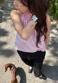 Parte de cima sem mangas em rosa claro com decote drapeado, calças largas pretas e sapatilhas pretas. Cão castanho passeando perto de um caminho.