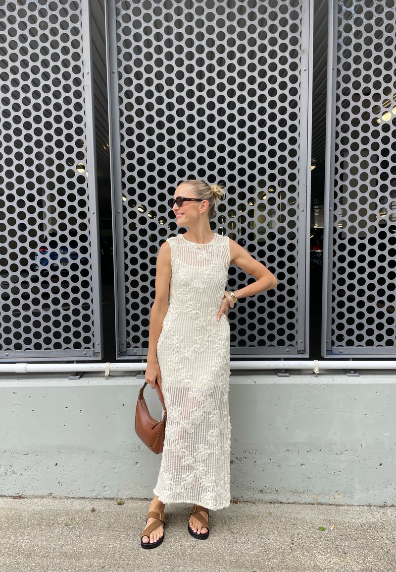 Vestito bianco a coste con motivi floreali, senza maniche, abbinato a una borsa marrone e sandali piatti, davanti a un muro perforato.