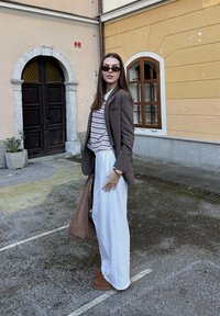 Rudas blazeris su juostuota marškinėliais, balti plačiakurčiai kelnių, ir rudi šlepetės. Priedai apima rudą rankinę ir saulės akinius.
