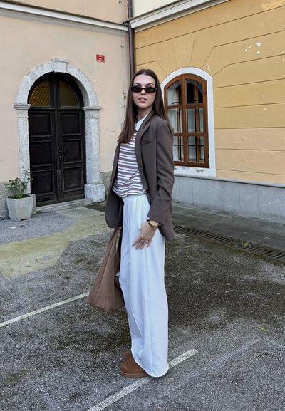 Blazer marrón sobre una camisa a rayas, pantalones anchos blancos y chanclas marrones. Los accesorios incluyen un bolso marrón y gafas de sol.