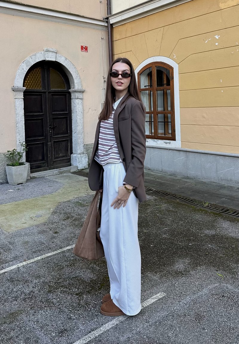 Brauner Blazer über einem gestreiften Hemd, weiße weit geschnittene Hosen und braune Slipper. Zu den Accessoires gehören eine braune Tasche und Sonnenbrillen.
