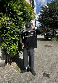 Fekete-fehér Adidas mez hosszú ujjakkal, feketére vágott nadrág, fekete sportcipő és fekete sapka egy zöld levél háttér előtt.