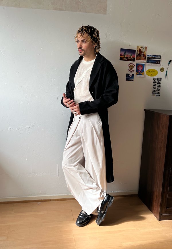 Persona con cabello rizado que lleva abrigo negro, suéter blanco, pantalones blancos anchos y zapatos negros apoyada contra una pared blanca con postales.