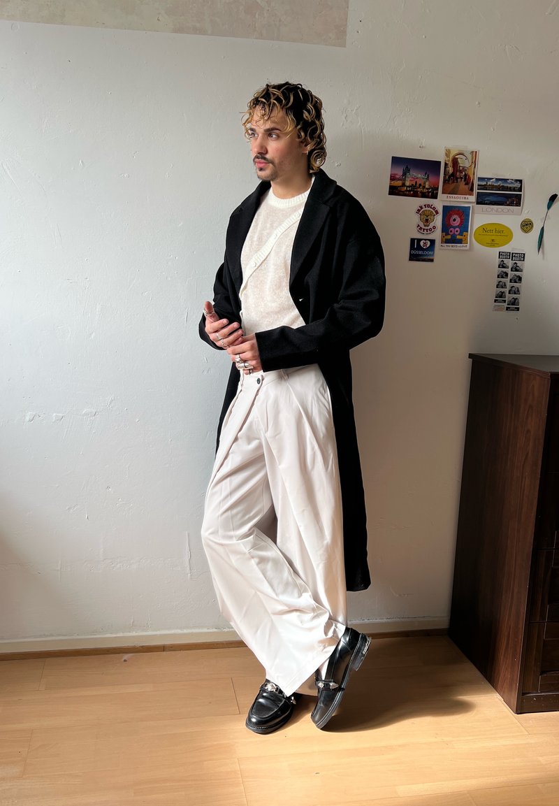 Personne aux cheveux bouclés portant un manteau noir, un pull blanc, un pantalon blanc large et des chaussures noires, appuyée contre un mur blanc avec des cartes postales.