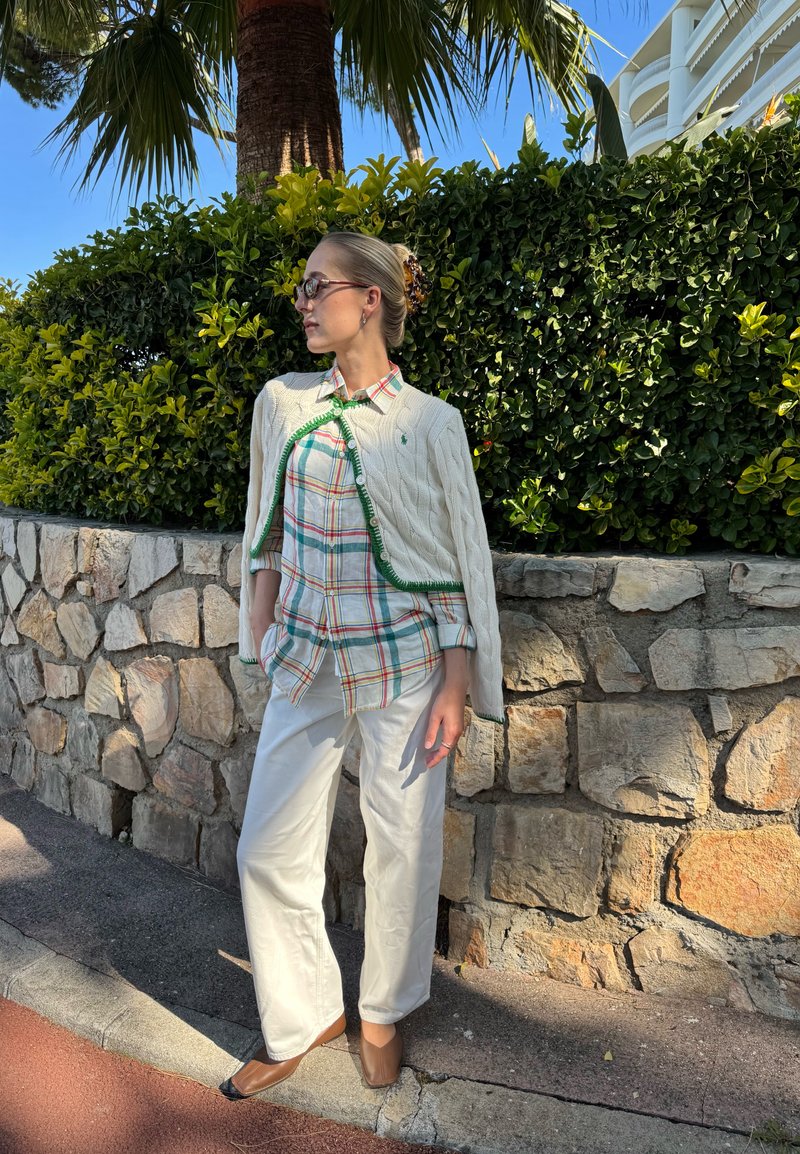 Femme portant des lunettes de soleil, un pull blanc à maille torsadée, une chemise à carreaux, un pantalon blanc et des chaussures marron, debout à côté d’un mur en pierre avec une haie verte.